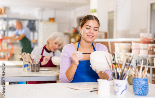 In pottery workshop, dark-haired woman paints vase that she made herself