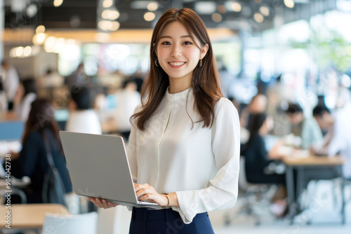 Smiling Japanese businesswoman standing in an office, holding her laptop. She is wearing casual clothes, and other people working behind her are blurred.,generated ai.