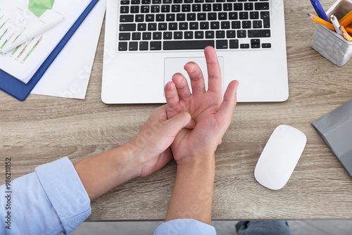 Man suffering from pain in wrist while working on laptop at wooden table, closeup, top view. Carpal tunnel syndrome