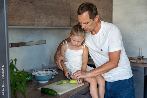 Father patiently teaching a his daughter how to cook in their home kitchen, family with dad and daughter preparing green salad with vegetables 