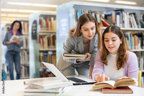 Two young women preparing to exam in library together, working on laptop computer, reading books and making notes