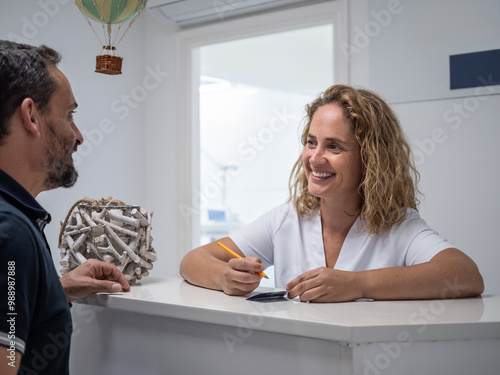 A friendly exchange between a receptionist and a patient takes place at a reception desk of a dental clinic, creating a welcoming atmosphere in a bright setting.