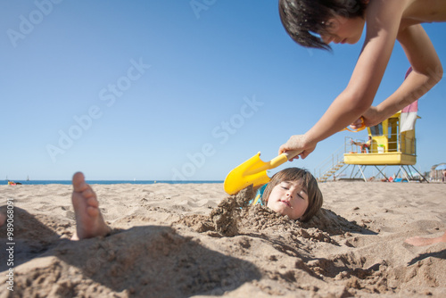 Boy is burying his brother under the sand on the beach.