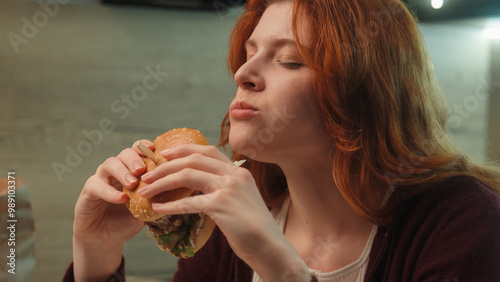 Young Caucasian woman female girl customer client eating junk fast food bite chewing burger hamburger cheeseburger eat pleasure diet cheat meal temptation indoors. Unhealthy snack overeating calories