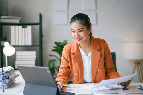 Young asian businesswoman is working at her desk in the office, smiling as she uses her tablet computer to go over paperwork