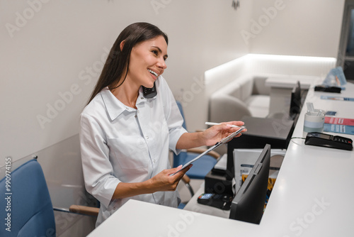 Young female medical receptionist practitioner nurse talking on phone with customers patients and making doctor appointment and writing on clipboard notebook at desk in hospital