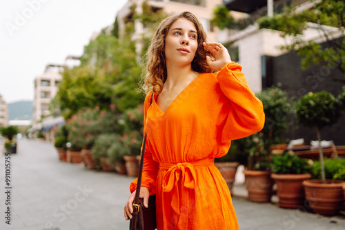 A woman wearing a vibrant orange dress strolls through an elegant city plaza, thoughtfully tucking her hair behind her ear as she admires the surroundings. Concept of lifestyle, fashion, travel.