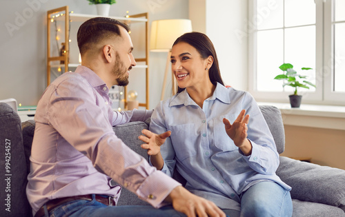 Happy smiling young woman telling something to her husband with man listening to her carefully. Loving couple sitting on sofa in living room, looking at each other, sharing feelings and emotions.