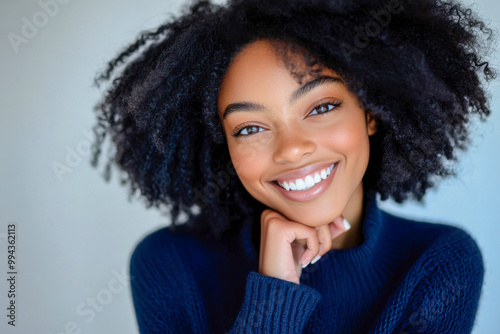 A woman with curly hair smiles brightly while resting her chin on her hand, showcasing natural beauty in a cozy indoor setting