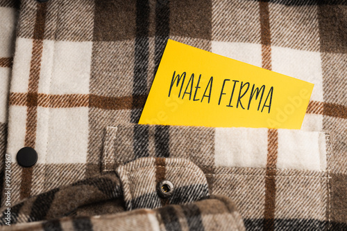 Yellow card with a handwritten inscription "Mała Firma", protruding from a brown plaid shirt (selective focus)