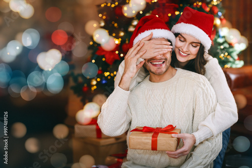 Loving young woman giving surprise gift to her husband, closing his eyes, celebrating Christmas at home