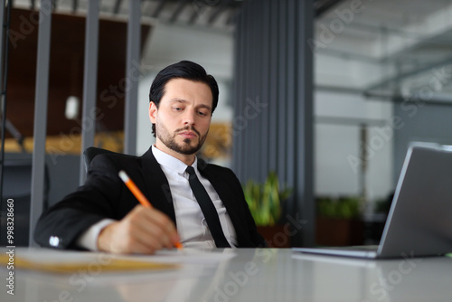 A man in a suit is writing with a pencil on a desk. Concept of professionalism and focus, as the man is likely working on a task or project. The presence of the laptop