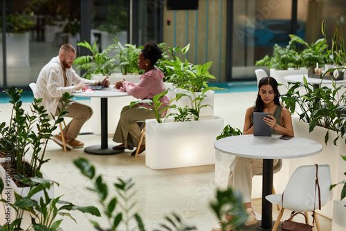 Wide angle shot of woman using tablet while sitting at table in cafe of office building. Business people working in cozy cafe featuring modern biophilic design