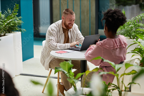 Full length shot of smiling adult man using laptop sitting at cafe table in office center while having work related conversation with female colleague, copy space