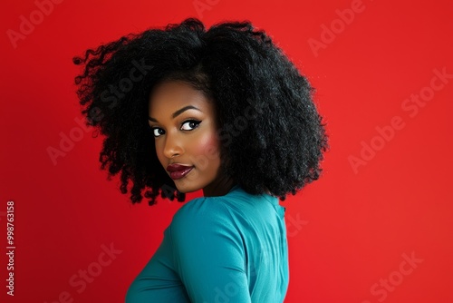 Confident Black woman with natural afro hairstyle in teal blue attire looking over her shoulder against a vibrant red background exuding cool and stylish vibes for diverse usage.