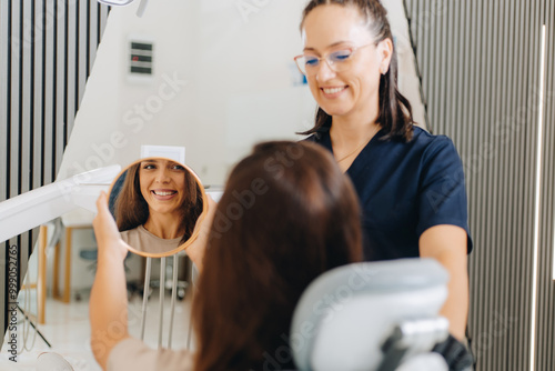 A patient sits in a dental chair, holding a mirror to see their smile while a dentist stands beside her, both smiling. The setting is a modern dental clinic with bright lighting.