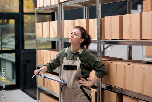 Delivery service employee searching clients parcel in warehouse checking clipboard with online order. Storage room worker wearing uniform overalls supervising goods in factory storehouse