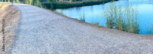 A gravel path runs alongside the serene waters of Calavera Lake in Carlsbad, California, with lush vegetation on the far side under a clear blue sky, creating a peaceful and scenic view.