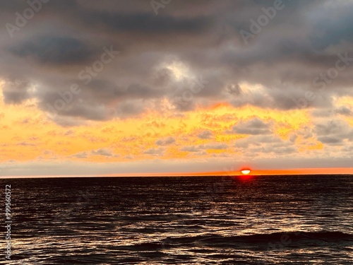 A stunning sunset over the ocean in Carlsbad, California, with vibrant orange clouds and the sun partially dipped below the horizon, reflecting on the water�s surface.