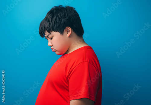 A side profile photograph of an overweight teen boy wearing a red t-shirt, standing with his head down, looking sad and dejected against a blue background