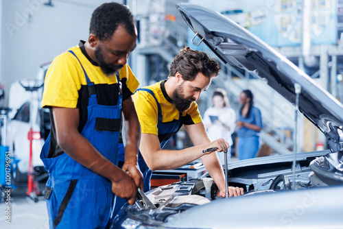 Certified servicemen in auto repair shop working together on fixing car, discussing best options. BIPOC worker and colleague collaborating on servicing broken vehicle, checking for faulty components