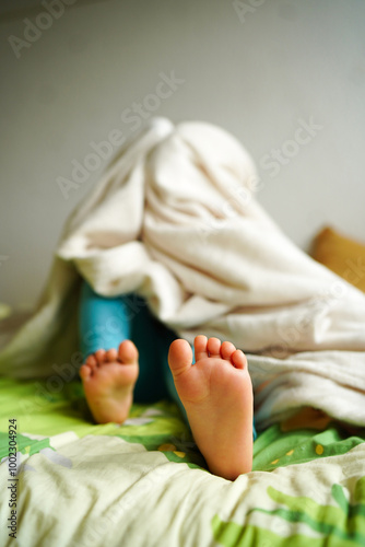 A young child hides under a blanket with bare feet sticking out, creating a playful and cozy scene on a bed. 