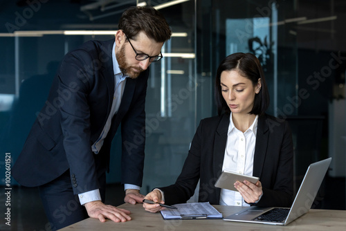 Business professionals engaged in workplace discussion over project details. Woman uses tablet for reference. Man leans over table, emphasizing teamwork. Office environment highlights technology