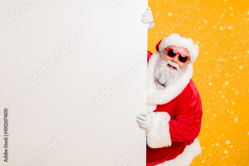 Portrait of funny funky christmas father in red hat headwear look behind white wall poster advertise winter x-mas shopping seasonal miracle sales discounts isolated over bright color background