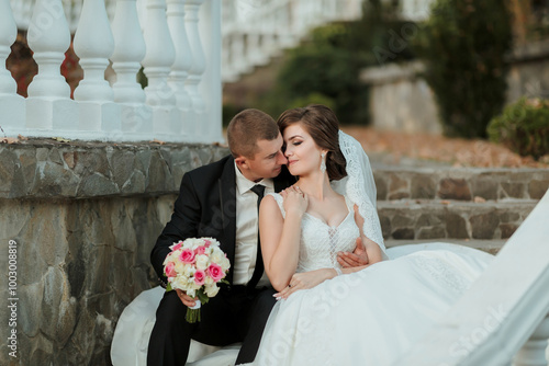 A bride and groom are sitting on a bench, the bride holding a bouquet of flowers. Scene is romantic and intimate