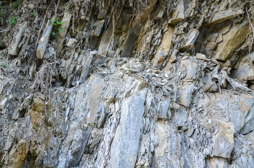 Grey rocky cliff face with visible layers and strata indicating geological processes, showcasing various sizes and shapes of stones and boulders. Texture typical of sedimentary rock formations