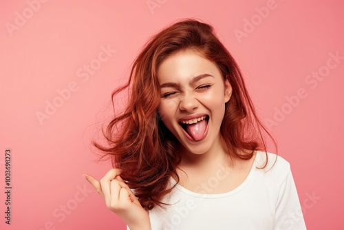 Young woman smiling with tongue out, winking at camera. Isolated on bright pink background. Happy, cheerful, caucasian girl with casual attire. Expressing joy, positive emotions.