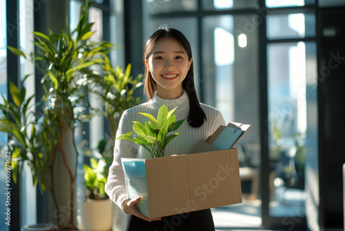 New employee starting new job holding cardboard box with belongings and plant smiling in modern office