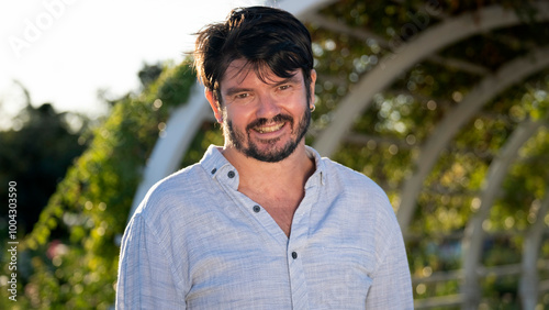 A middle-aged, dark-haired businessman wearing a shirt is smiling at the camera in a sunny, tree-filled park on a summer day