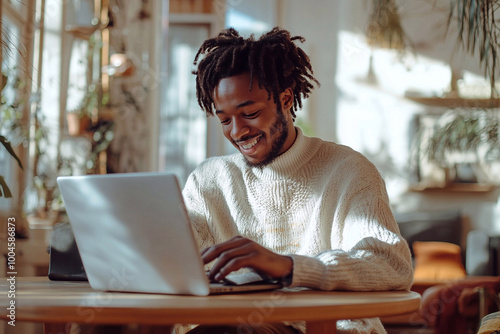 A young man of African descent sits at a wooden table and works on a laptop