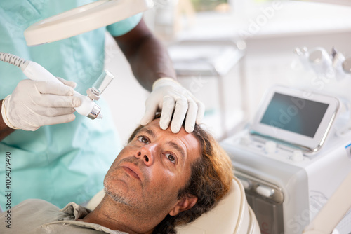 During visit to cosmetology office, client man undergoes facial skin moisturizing procedure using water-oxygen sprayer to smooth dermis and saturate deep layers of skin with moisture