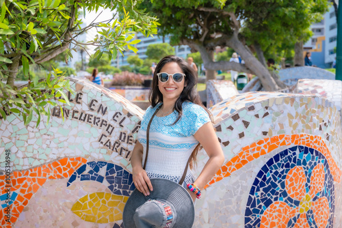 Young Latina woman smiling and sitting on a colorful mosaic bench in Love Park, Miraflores, Lima, Peru, enjoying a sunny day.