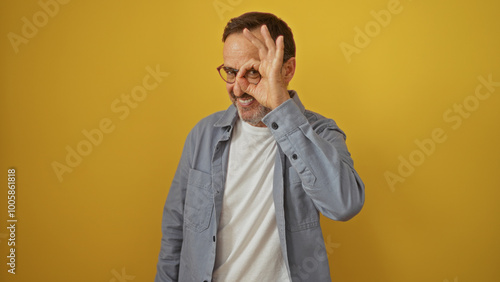 Hispanic man making ok gesture with hand in front of eye, in front of an isolated yellow background, smiling and looking cheerful.