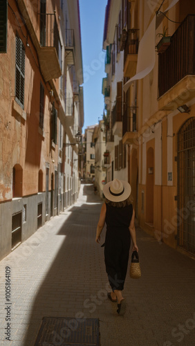 Woman walking through a narrow street in palma, mallorca, spain, wearing a hat and holding a wicker bag, under a clear blue sky in an outdoor setting surrounded by old buildings