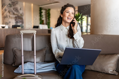 A woman sits in a hotel lobby using a laptop and talking over a smartphone while her suitcase stands nearby. She appears focused on her work in a comfortable, modern lounge area.