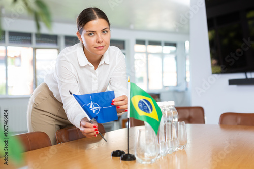 Positive young woman putting little flag of NATO on table next to the flag of Brazil and bottles of water in conference room