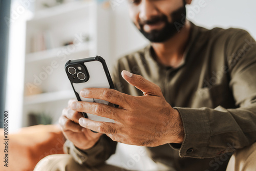 Young man smiling while holding mobile phone, using mobile app at home, selective focus on hand