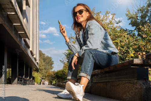 City woman lifestyle. Brunette stylish woman in denim jacket and sunglasses sitting on bench on city street using mobile phone and smiling