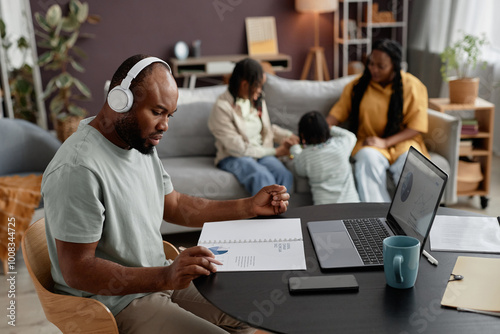 Man wearing headphones concentrating on work with laptop while family gathering in background interacting and playing with child in a cozy living room setting