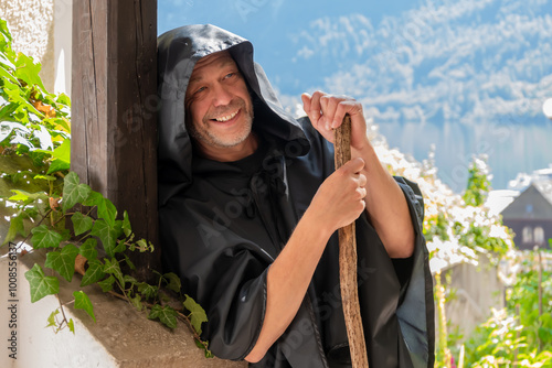 A cheerful monk in a black cassock and carrying a staff leaned against the wooden railing of a staircase against a backdrop of mountains.