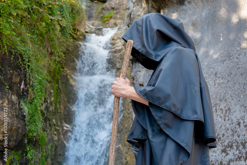 A monk in a black robe with a staff walks past a mountain waterfall.