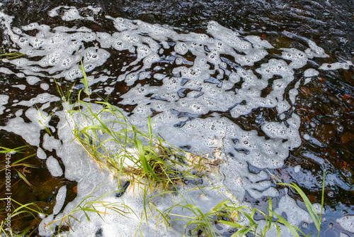 River with white foam caused by pollution or sewage runoff into water with grass at edge of stream