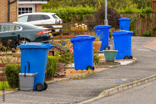 Blue waste recycling wheelie bins trash cans lined up in a row for collection on a residential street with parked cars in driveways