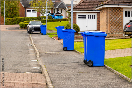 Blue waste recycling wheelie bins trash cans lined up in a row for collection on a residential street with parked cars