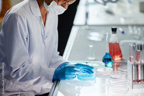 Cropped shot of female microbiologist in medical gloves sitting at workstation observing bacterial colony in petri dish while dripping liquid from pipette in laboratory, copy space