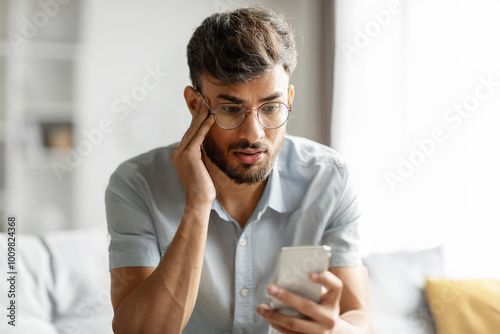 Shocked indian guy looking at cellphone screen, sitting on couch at home, holding phone and touching his head, reading bad news or message, copy space